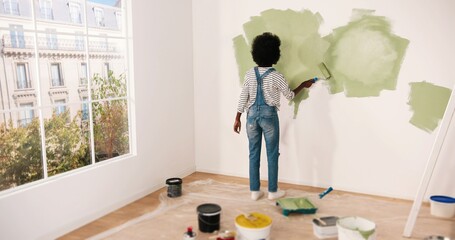 Young African American female alone painting wall with roller brush in olive color while renovating...