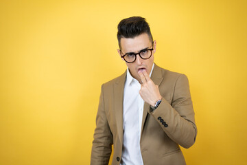 Young business man over isolated yellow background disgusted with hand inside his mouth