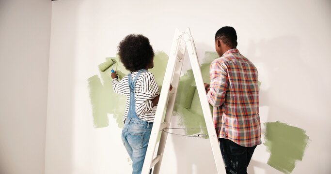 Back View Of African American Couple Male And Female Redesigning And Renovating Room In Own Apartment Standing On Ladder Painting Walls In Green Olive Color With Roller Brush. Home Repair Concept.