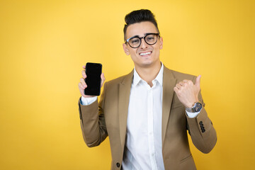 Young business man over isolated yellow background smiling, showing the phone and pointing side