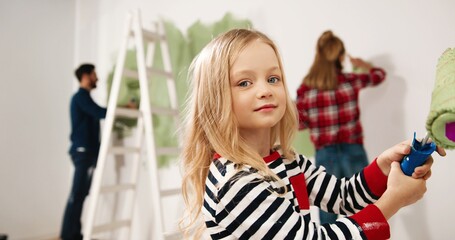 Close up of happy smiling cute little girl painting wall with roller brush in hands helping her Caucasian young parents to renovate house. Mom and dad working and waving hands on background