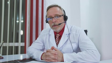 Serious doctor giving audio consultation call. Professional senior therapist sitting in medical office with headset and talking. Remote consultation.