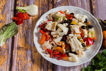 cod boiled with winter cherry tomatoes, black olives, celery, onion, pickled peppers on wooden background. Selective focus.