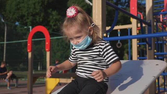 little girl wears a medical safety mask on his face. kid is playing outside in children's park. concept of kid safety from contracting an infection. girl is protected in a safety mask in park.