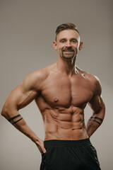 A smiling muscular man with a beard is posing. The athletic guy is squeezing his muscles. A bodybuilder with tattoos on his forearms is standing with his hands on his hips