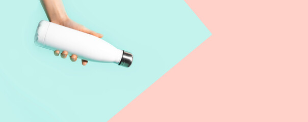 Close-up of female hand holding reusable thermo bottle. Backgrounds of pastel blue and coral pink colors. Panoramic photo with copy space.