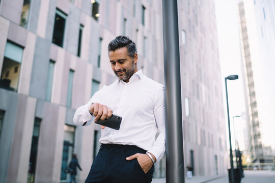 Caucasian businessman in white shirt waiting corporate partner in financial downtown checking time of meeting on wearable wrist watch, confident male proud CEO with cellphone looking on smartwatch
