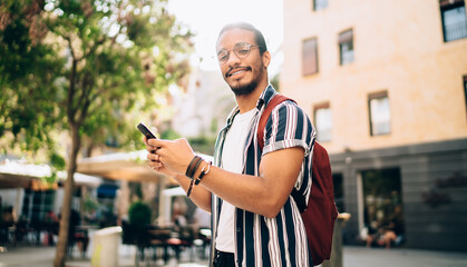 Portrait of cheerful dark skinned male traveler using smartphone for navigation during sunny day in town, smiling african american hipster guy sending messages in online chat spending time outdoors