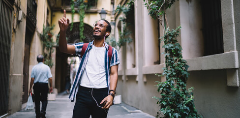 Joyful ethnic man walking in old town and using smartphone with audioguide