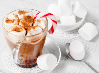 a transparent cup of cocoa drink with marshmallows and candy cane on a whitish wooden table, beside a spoon and other marshmallows