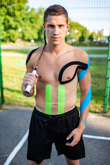 Portrait of handsome young caucasian professional tennis player with elastic tapes on body carrying racket near fenced sports ground. Confident man with muscular naked torso looking at camera.