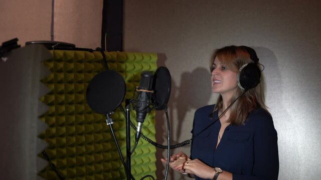 A woman in a small recording studio sings into a microphone wearing black headphones in front of a studio microphone. A woman sings a song in a recording studio.