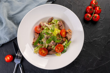 salad with meat and cherry tomatoes in a white plate on a dark background