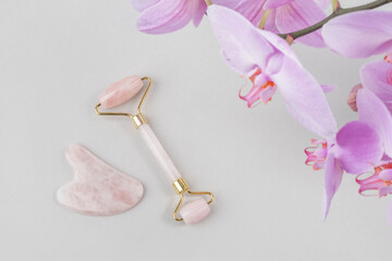 Crystal rose quartz facial roller, massage tool jade Gua sha and natural orchid flower on grey background. Facial anti-age massage for natural lifting and toning treatment at home