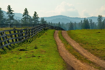 Russia. South Of Western Siberia, Mountain Altai. Green pastures in the mountain valleys fenced in by a wooden fence along the dirt roads.
