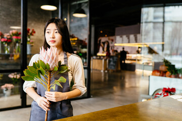 Creative millennial female designer, florist holding pink protea in modern interior of flower shop. Selling plants, bouquets, floral composition, small business concept. Copy space for text.