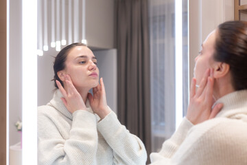 Obraz na płótnie Canvas Young beautiful woman in a sweater in a beauty salon looks in the mirror, touches her face, thinks about the upcoming procedures, considers herself