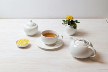still life with tea in cup with saucer, teapot, sugar bowl, yellow rose in milk jug and lemon on white background