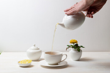 woman hands pouring tea in cup with saucer from tea pot, sugar bowl, yellow rose in milk jug and lemon on white background