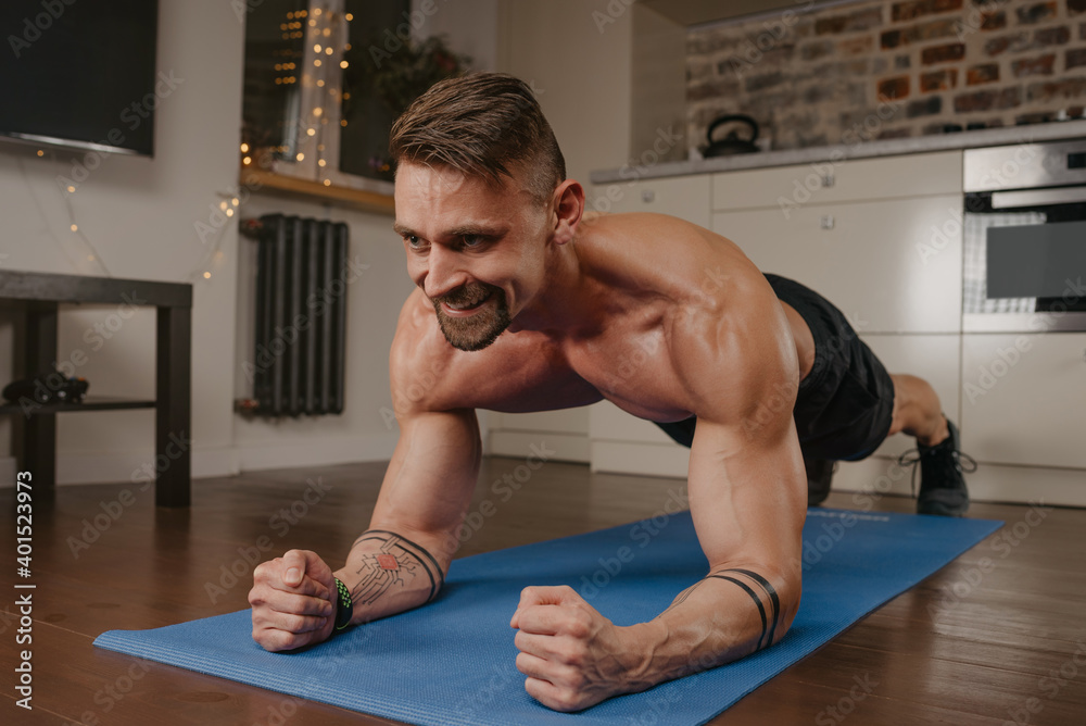 Wall mural A happy muscular man with a naked torso is doing a plank on a blue yoga mat in his apartment in the evening. A smiling bodybuilder with tattoos on his forearms is training at home.