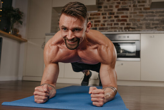 A muscular man with a naked torso is doing a plank on a blue yoga mat in his apartment in the evening. A smiling athletic guy with tattoos on his forearms is training at home. - Powered by Adobe