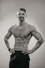 A black and white photo of a muscular man with a beard who is posing. The athletic guy is demonstrating his sporty physique. A bodybuilder with tattoos on his forearms is relaxing after hard training.