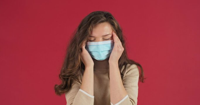 Portrait of female sad face portrait in studio on red background young woman in protective medical mask feels headache suffers from migraine high temperature holds her head with hands disease concept