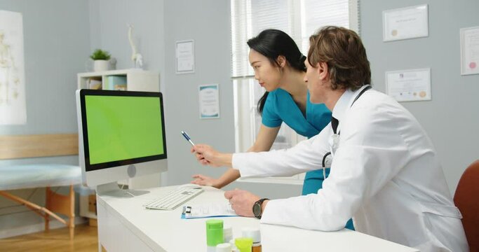 Side view. Portrait of Caucasian male doctor sitting in cabinet in hospital and looking at computer green screen, chatting with Asian female nurse explaining diagnosis and treatment chroma key concept