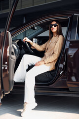 Sits with car's window opened. Fashionable beautiful young woman and her modern automobile