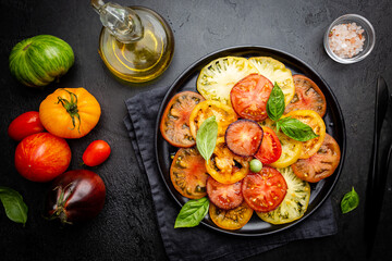 Ripe fresh colorful tomatoes salad with olive oil and basil leaves on black background, top view