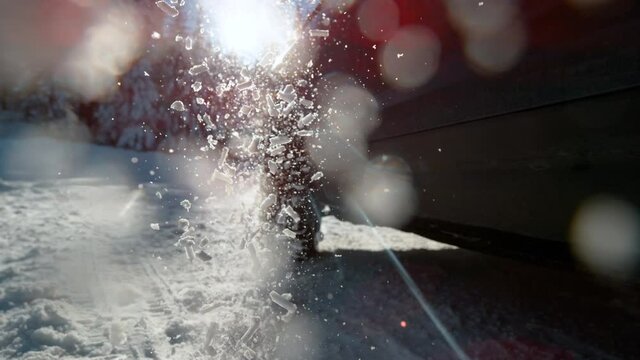 SLOW MOTION, LENS FLARE, CLOSE UP, DOF: Pieces of snow glisten in the bright winter sunshine as they fly in the air from spinning wheels of a car having difficulties getting traction on an icy road.
