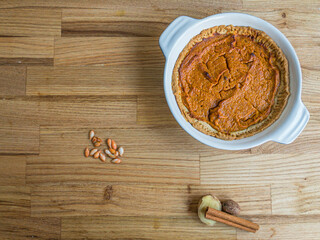 Homemade butternut squash pie with pumpkin spice, next to a cinnamon stick, piece of ginger nutmeg and a few squash seeds on a wooden surface with copy space