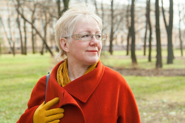 Mature blonde shorthaired woman  in red coat, golden glasses and yelllow glowes in spring park