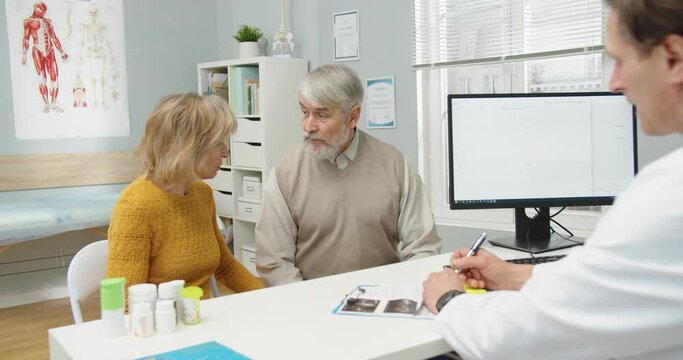 Rear of male physician sits in hospital office and talking to old patients about disease and treatment Caucasian senior wife and husband at doctor appointment in clinic cabinet. Healthcare concept