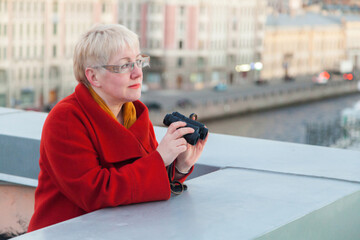 A mature woman in glasses with short blond hair in red coat stands with binoculars on the roof of an old house in St. Petersburg