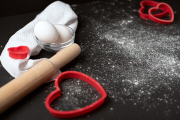 Obraz na płótnie Canvas Red heart-shaped baking tins for cookies and wooden rolling pin with scattered flour on the dark (black) background. Flat lay, top view, space for text. Valentine's day concept.