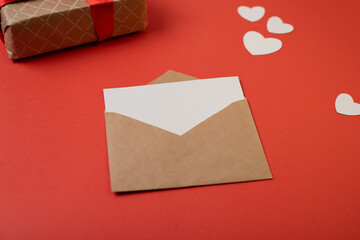 Craft envelope with blank white note mockup inside and Valentines hearts on red background. Flat lay, top view. Romantic love letter for Valentine's day concept.