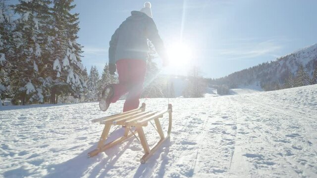 SLOW MOTION, CLOSE UP, LENS FLARE, LOW ANGLE: Unrecognizable young woman runs along a snowy meadow and pulling her sled on a sunny winter day. Female tourist is having fun sleighing in Kranjska Gora.