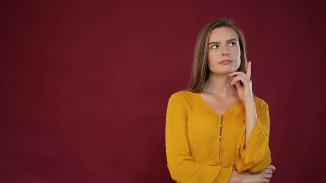 A pensive, doubting woman in a blouse ponders and imagines in her mind, wanders around a difficult decision, feels confusion, unsure of her choice, throws up her hands against a red background.