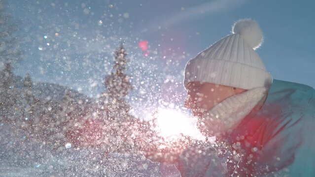 SLOW MOTION TIME WARP, LENS FLARE, CLOSE UP, DOF: Smiling young Caucasian woman blows away a handful of fresh powder snow. Playful female tourist blowing snowflakes into the bright winter sunshine.