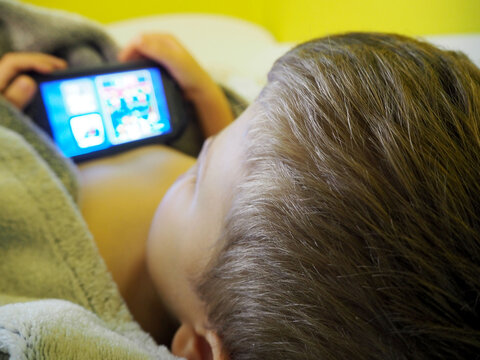 A Blond Boy Of Five Years Old Lies In Bed Under A Gray Blanket And Looks Into A Black Smartphone Rear View