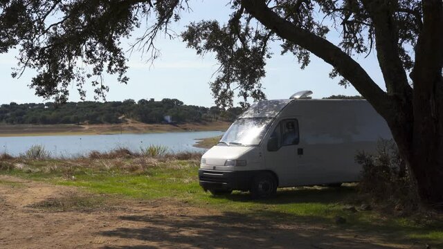 Camper van on an Alentejo dam landscape with the lake behind and under the shade of a tree, in Portugal