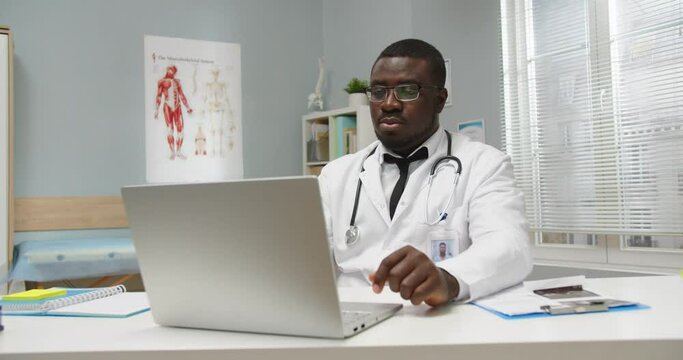 Portrait of handsome young serious African American general practitioner doctor sitting at table in cabinet in medical center working, typing and searching internet on laptop. Health concept