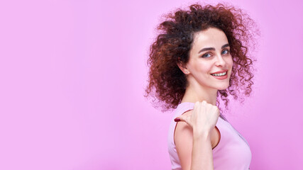 Portrait of smiling caucasian curly girl points finger to the side on copy space isolated in pink studio background