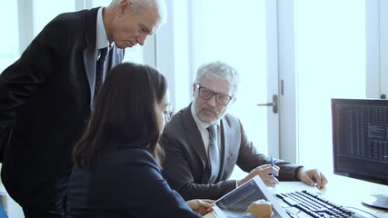 Confident female finance professional explaining charts on papers to two mature businessmen, pointing at graphs and monitor and speaking. Business meeting or analysis concept