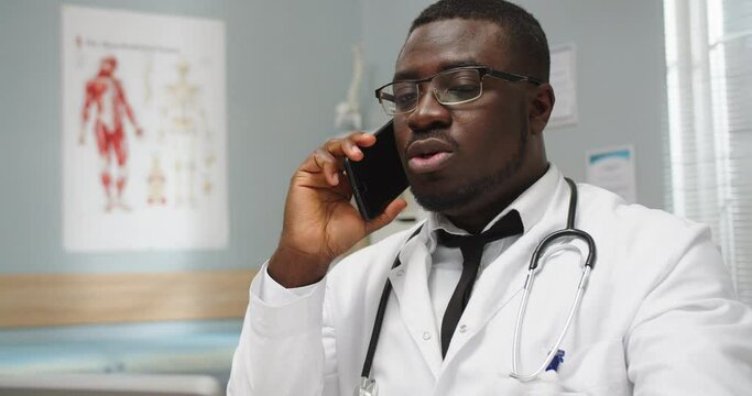 Young serious African American general practitioner physician sitting in cabinet in medical center calling on smartphone, talking on cellphone. Healthcare. Consulting over phone. Close up concept