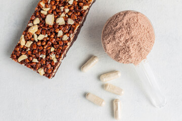 Scoop of whey or soy protein isolate, chocolate protein bar, white capsules of amino acids and creatine, bodybuilding food supplements on white background, top view, flat lay