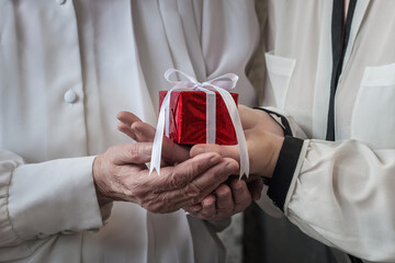 Heartwarming moment of life with closeup on hands. Young woman in white blouse giving a gift box wrapped in red shining paper and white ribbon to eldery woman (mother or granny) for event or holiday
