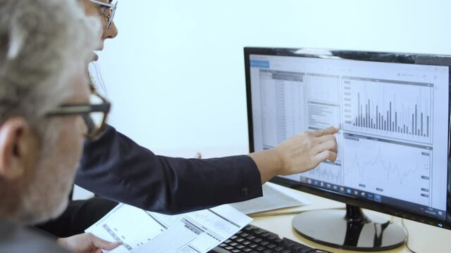 Serious female finance professional explaining data and charts to two mature businessmen, pointing at monitor and speaking. Business meeting or marketing concept