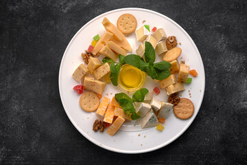 Cheese plate, assorted cheeses with mint, candied fruit, honey and cookies, on a white plate, on a dark background